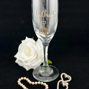 May include: A champagne flute with the word "Dulhan" in gold script, a gold lotus flower design, and a small heart. A white rose and two pearl bracelets are also in the image.