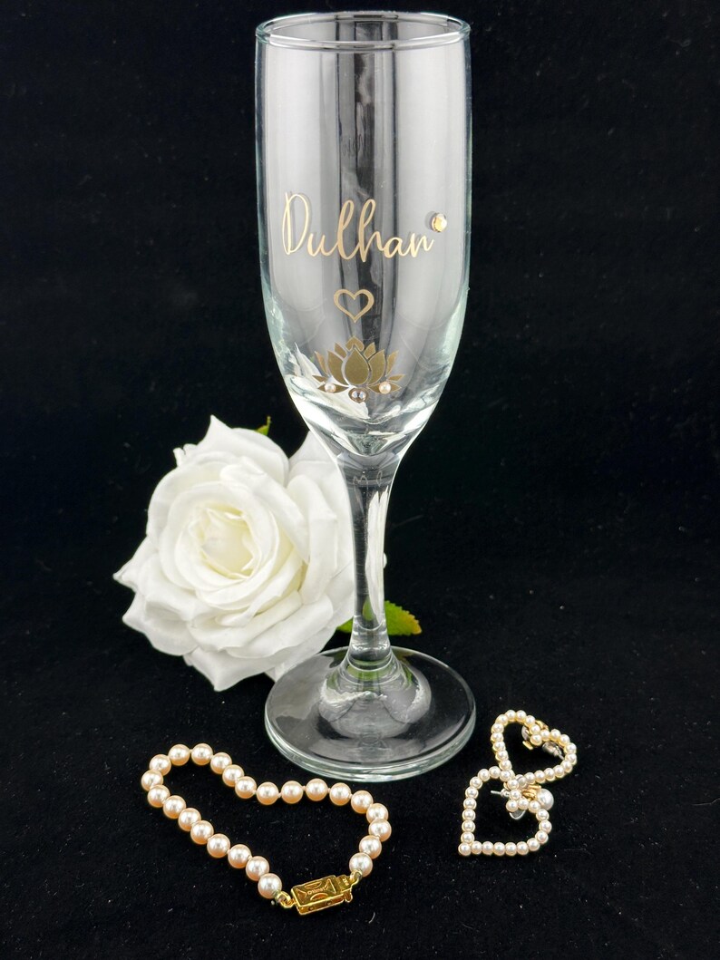 May include: A champagne flute with the word "Dulhan" in gold script, a gold lotus flower design, and a small heart. A white rose and two pearl bracelets are also in the image.