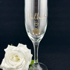 May include: A clear champagne flute with gold lettering that reads "Dulhan", a heart, and a lotus flower design. The flute has a long stem and sits on a black surface. A white rose is in the foreground.