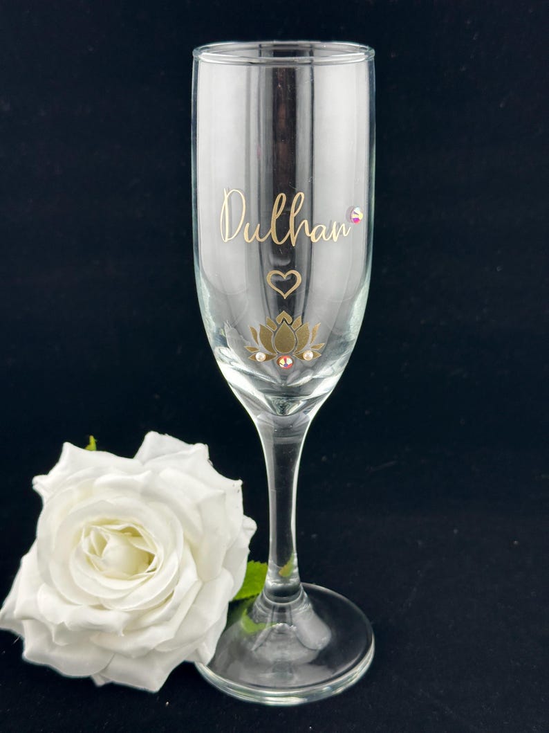 May include: A clear champagne flute with gold lettering that reads "Dulhan", a heart, and a lotus flower design. The flute has a long stem and sits on a black surface. A white rose is in the foreground.