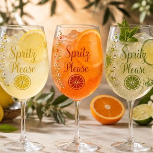 May include: Three clear glass wine glasses filled with colorful cocktails, ice, and fruit slices. Each glass features the text Spritz Please, a citrus fruit icon, and a vertical line of small decorative rhinestones along the side of the bowl.