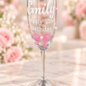 May include: A clear glass champagne flute featuring white text that reads Emily Happy 21st Birthday. The glass is decorated with a pink bow graphic and scattered white and pink pearl-like beads attached to the exterior surface of the glass.