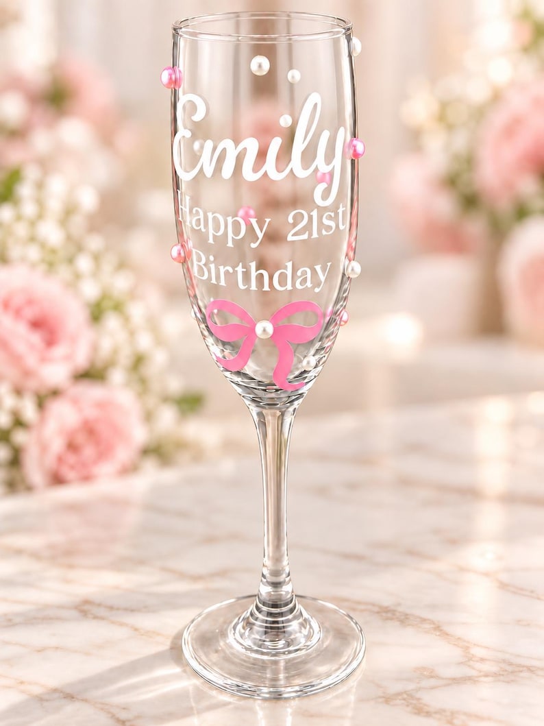 May include: A clear glass champagne flute featuring white text that reads Emily Happy 21st Birthday. The glass is decorated with a pink bow graphic and scattered white and pink pearl-like beads attached to the exterior surface of the glass.