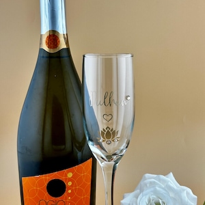 May include: A bottle of Ogio Prosecco DOC Italia next to a champagne flute with the word "Mulher" and a lotus flower design. A white rose is on the right. The bottle has an orange label.