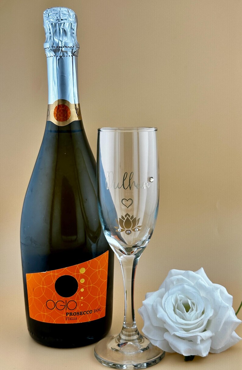 May include: A bottle of Ogio Prosecco DOC Italia next to a champagne flute with the word "Mulher" and a lotus flower design. A white rose is on the right. The bottle has an orange label.