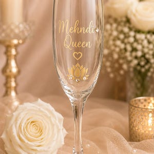 May include: A clear champagne flute with gold lettering that reads "Mehndi Queen", a heart, and a lotus flower design. The glass is decorated with small, pearl-like embellishments. The background includes a lit candle and white roses.