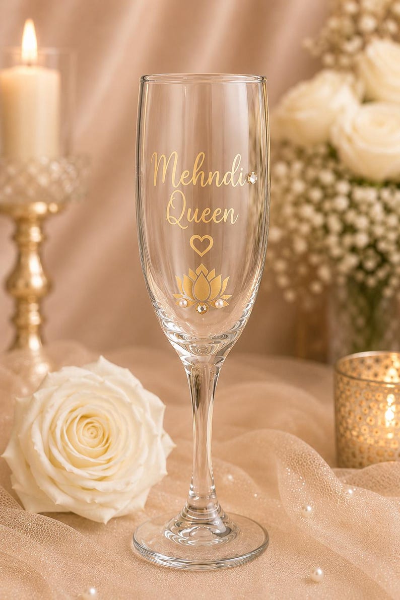 May include: A clear champagne flute with gold lettering that reads "Mehndi Queen", a heart, and a lotus flower design. The glass is decorated with small, pearl-like embellishments. The background includes a lit candle and white roses.