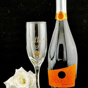 May include: A champagne bottle and a personalized champagne flute with the name "Dulhan" in gold script, a heart, and a lotus flower design. A white rose and a pearl bracelet are also in the image. The bottle has an orange label.