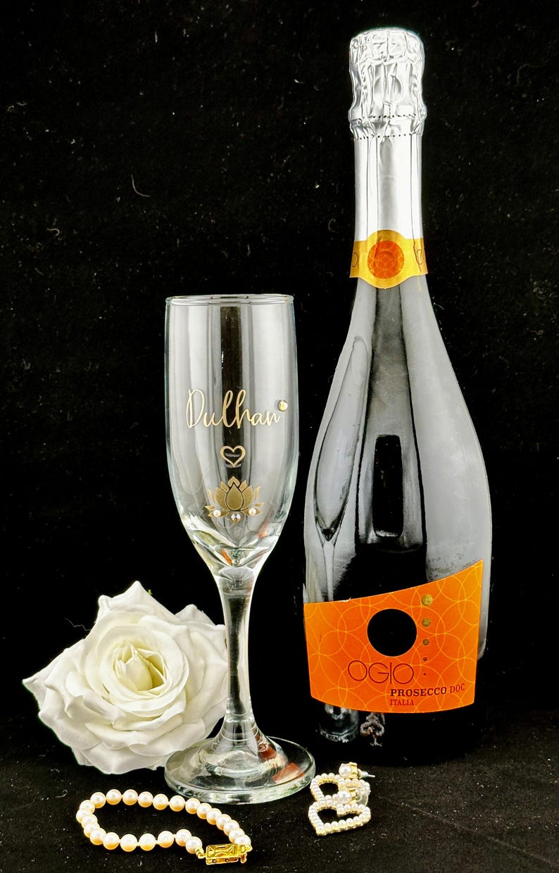 May include: A champagne bottle and a personalized champagne flute with the name "Dulhan" in gold script, a heart, and a lotus flower design. A white rose and a pearl bracelet are also in the image. The bottle has an orange label.