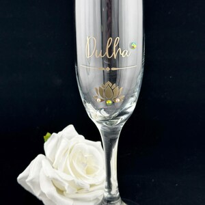 May include: A clear champagne flute with the word "Dulha" in gold script, a gold lotus flower design, and decorative gems. A white rose rests near the base of the glass. The background is black.