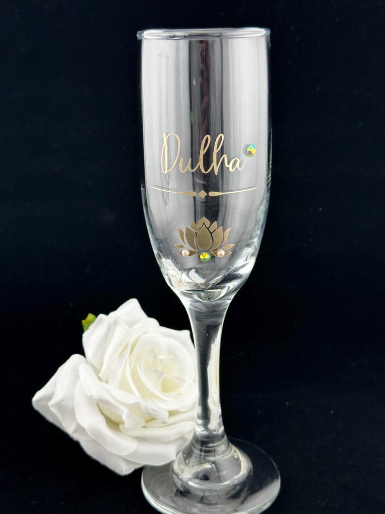 May include: A clear champagne flute with the word "Dulha" in gold script, a gold lotus flower design, and decorative gems. A white rose rests near the base of the glass. The background is black.