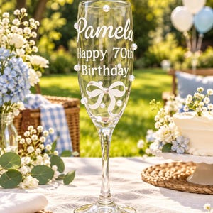 May include: A clear glass champagne flute decorated with white pearls and a white bow graphic. The text "Pamela" is printed in a script font above the text "Happy 70th Birthday" in a sans-serif font. The glass has a tall, slender stem.