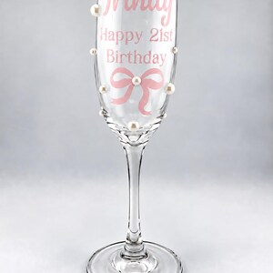 May include: A clear glass champagne flute decorated with white pearls and pink text that reads Trinity, Happy 21st Birthday, and a pink bow graphic. The stem and base are made of clear glass, and the pearls are attached to the exterior of the bowl.