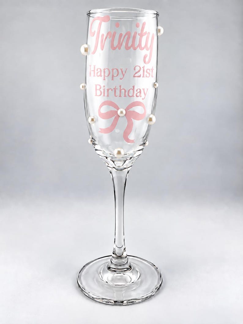 May include: A clear glass champagne flute decorated with white pearls and pink text that reads Trinity, Happy 21st Birthday, and a pink bow graphic. The stem and base are made of clear glass, and the pearls are attached to the exterior of the bowl.