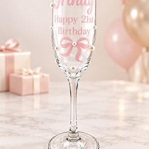 May include: A clear glass champagne flute decorated with white faux pearls and pink text that reads Trinity Happy 21st Birthday, featuring a pink ribbon graphic printed on the front surface of the glass bowl and a slender stem with a round base.