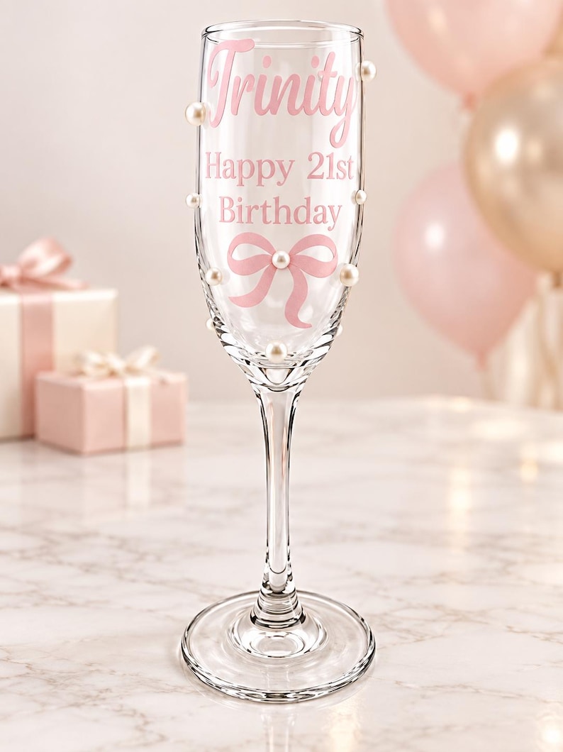 May include: A clear glass champagne flute decorated with white faux pearls and pink text that reads Trinity Happy 21st Birthday, featuring a pink ribbon graphic printed on the front surface of the glass bowl and a slender stem with a round base.