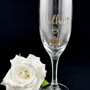 May include: A clear champagne flute with gold lettering that reads "Dulhan". The glass is decorated with a gold lotus flower, a heart, and pink gems. A white rose is in the background.