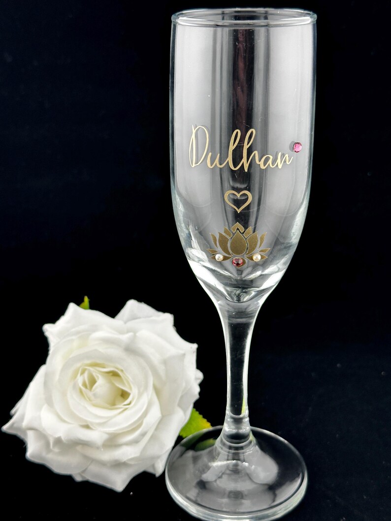 May include: A clear champagne flute with gold lettering that reads "Dulhan". The glass is decorated with a gold lotus flower, a heart, and pink gems. A white rose is in the background.