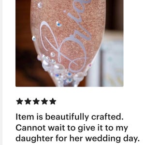 May include: A clear glass champagne flute filled with a sparkling pink beverage. The glass features the white cursive text Bride and is decorated with various sizes of white pearls and small iridescent rhinestones near the base of the bowl.