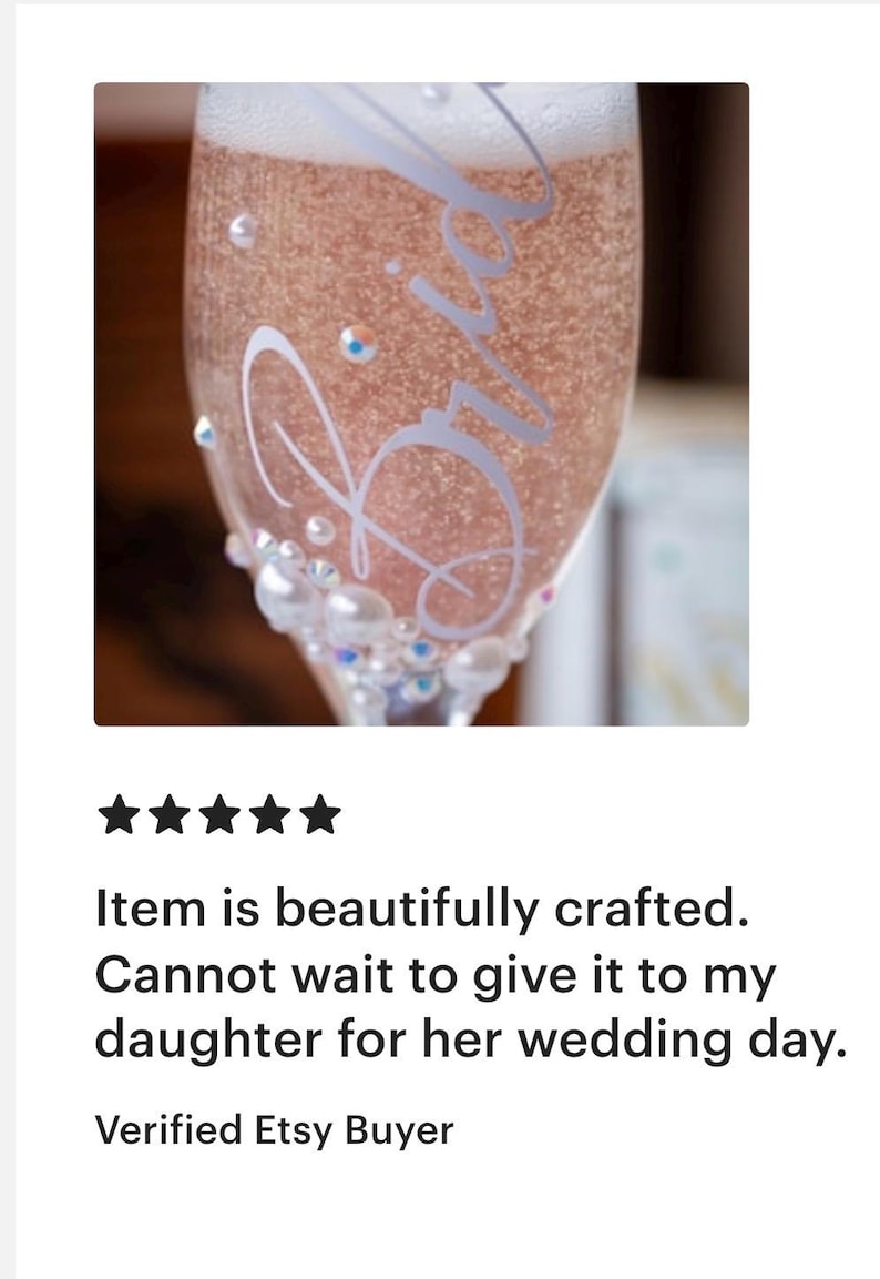 May include: A clear glass champagne flute filled with a sparkling pink beverage. The glass features the white cursive text Bride and is decorated with various sizes of white pearls and small iridescent rhinestones near the base of the bowl.