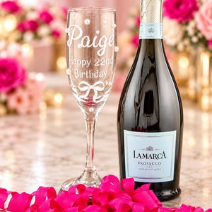 May include: A clear glass champagne flute featuring white script text that reads Paige Happy 22nd Birthday, accented with a white bow graphic and small white pearls, positioned next to a dark glass bottle of La Marca Prosecco sparkling wine.