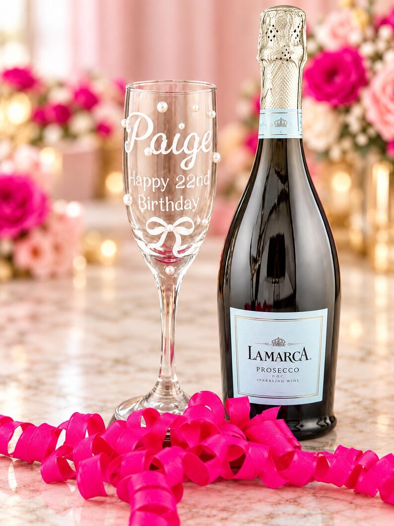 May include: A clear glass champagne flute featuring white script text that reads Paige Happy 22nd Birthday, accented with a white bow graphic and small white pearls, positioned next to a dark glass bottle of La Marca Prosecco sparkling wine.