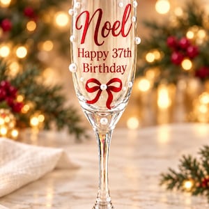 May include: A clear glass champagne flute decorated with white pearls, a red ribbon graphic, and red text that reads Noel Happy 37th Birthday. The stem and base are made of clear glass, and the bowl is adorned with scattered white pearl accents.
