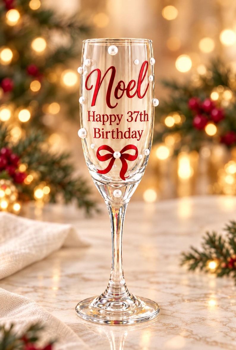 May include: A clear glass champagne flute decorated with white pearls, a red ribbon graphic, and red text that reads Noel Happy 37th Birthday. The stem and base are made of clear glass, and the bowl is adorned with scattered white pearl accents.