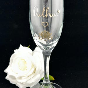 May include: A clear champagne flute with gold lettering that reads "Dulhan". The glass is decorated with a gold lotus flower, a heart, and small crystals. A white rose is in the background.
