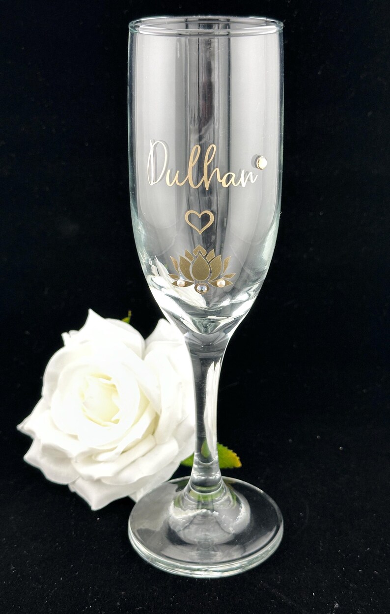 May include: A clear champagne flute with gold lettering that reads "Dulhan". The glass is decorated with a gold lotus flower, a heart, and small crystals. A white rose is in the background.