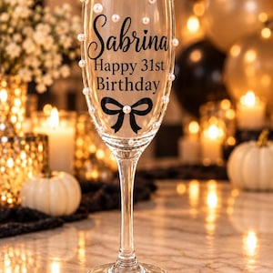 May include: A clear glass champagne flute featuring the black printed text Sabrina Happy 31st Birthday above a black bow graphic. The exterior of the glass is decorated with small white pearl-like beads scattered across the surface of the bowl.