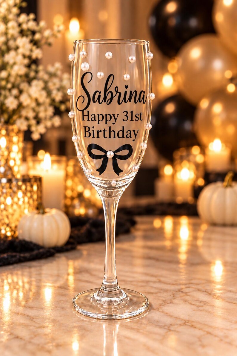 May include: A clear glass champagne flute featuring the black printed text Sabrina Happy 31st Birthday above a black bow graphic. The exterior of the glass is decorated with small white pearl-like beads scattered across the surface of the bowl.