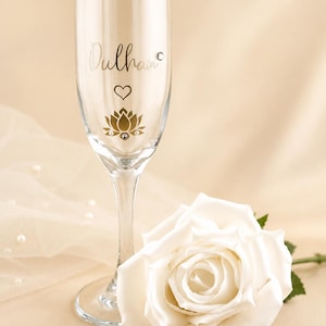 May include: A clear champagne flute with the word "Dulhan" in gold script, a heart, and a lotus flower design. A white rose lies next to the glass. The background is a soft, neutral color.