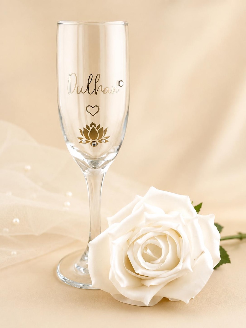 May include: A clear champagne flute with the word "Dulhan" in gold script, a heart, and a lotus flower design. A white rose lies next to the glass. The background is a soft, neutral color.