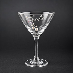 May include: A clear glass martini glass featuring the word Bride in white script lettering. The glass is decorated with a cascading arrangement of small white pearls and iridescent rhinestones that extend downward from the text.