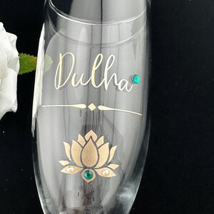May include: A clear champagne flute with the word "Dulha" in gold script. The glass features a gold lotus flower design with a green gem in the center and two pearls. A green gem is also placed near the top of the glass.