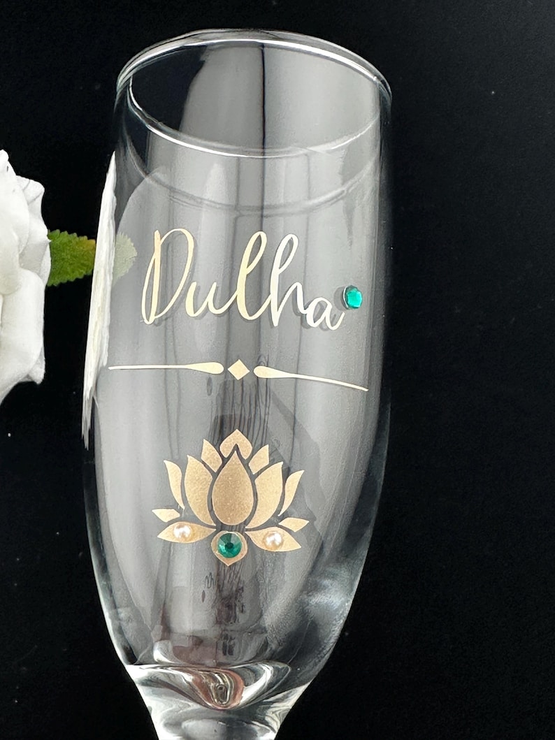 May include: A clear champagne flute with the word "Dulha" in gold script. The glass features a gold lotus flower design with a green gem in the center and two pearls. A green gem is also placed near the top of the glass.