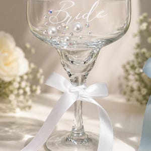 May include: A clear glass coupe with a wide bowl and a stem. The word Bride is printed in white script on the bowl, accented with white pearls and iridescent crystals. A white satin ribbon is tied in a bow around the stem of the glass.