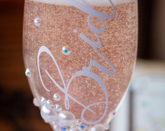 May include: A clear glass champagne flute filled with a sparkling pink beverage. The glass features the white cursive text Bride and is decorated at the base with an assortment of white pearls and small iridescent rhinestones of varying sizes.