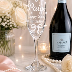 May include: A clear glass champagne flute featuring white text that reads Paige Happy 22nd Birthday, a white bow graphic, and several small white pearl embellishments adhered to the exterior surface of the glass bowl and stem.