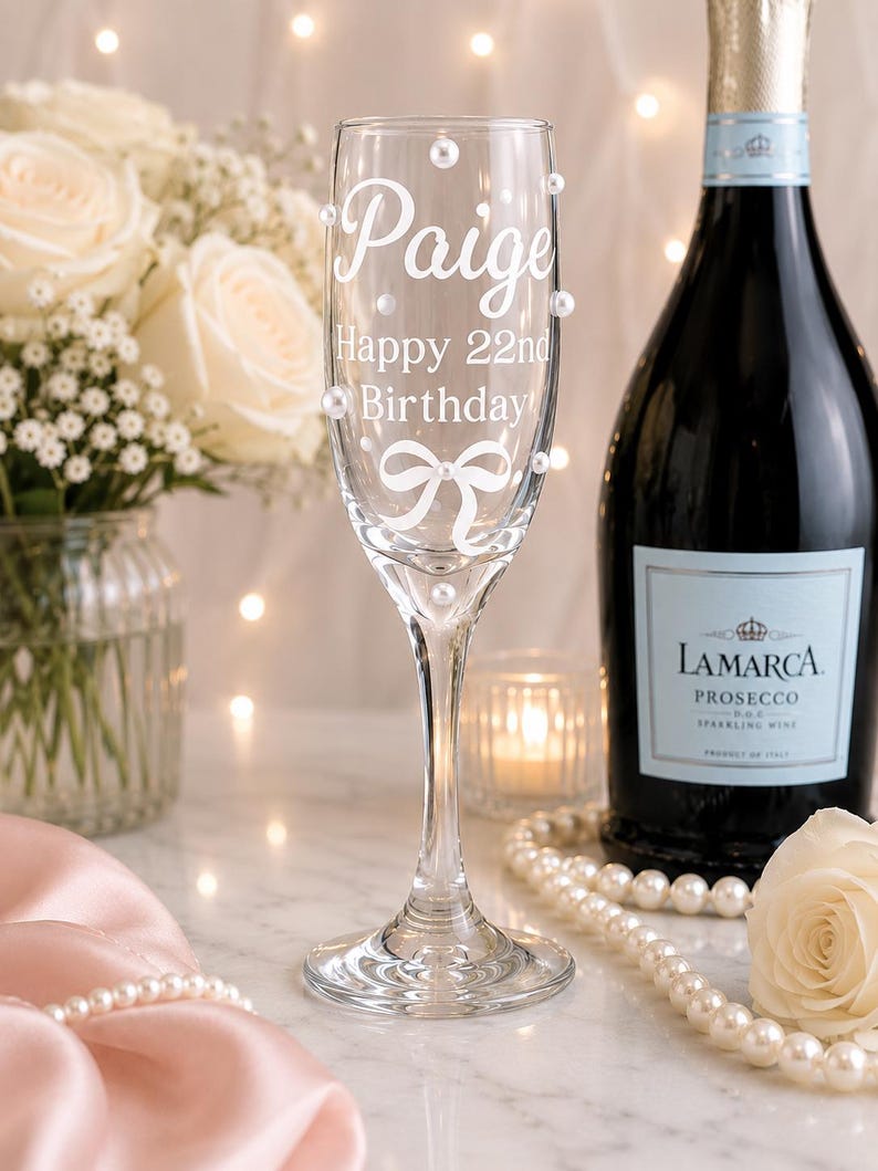May include: A clear glass champagne flute featuring white text that reads Paige Happy 22nd Birthday, a white bow graphic, and several small white pearl embellishments adhered to the exterior surface of the glass bowl and stem.