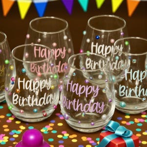 May include: A collection of clear stemless wine glasses, each featuring the text Happy Birthday in various colors and fonts. The glasses are decorated with scattered multicolored pearl-like beads and small rhinestones across the exterior surface.
