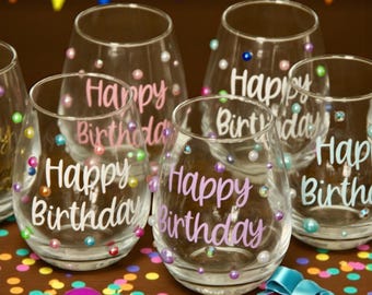May include: A collection of clear stemless wine glasses, each featuring the text Happy Birthday in various colors and fonts. The glasses are decorated with scattered multicolored pearl-like beads and small rhinestones across the exterior surface.