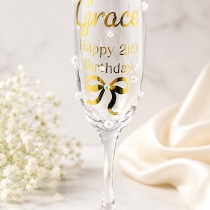 May include: A clear glass champagne flute decorated with gold lettering that reads Grace Happy 21st Birthday. The glass features a gold bow graphic with a central rhinestone and is embellished with scattered white pearl beads on the exterior surface.