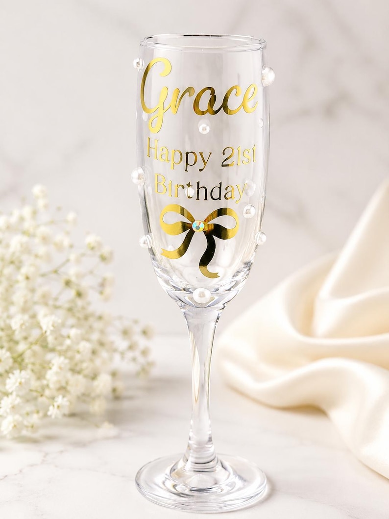 May include: A clear glass champagne flute decorated with gold lettering that reads Grace Happy 21st Birthday. The glass features a gold bow graphic with a central rhinestone and is embellished with scattered white pearl beads on the exterior surface.