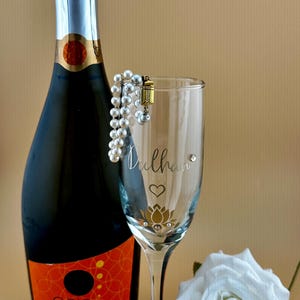 May include: A champagne bottle and a personalized champagne flute with the word "Dulhan" and a lotus flower design. A pearl bracelet is draped over the glass, and a white rose is in the background.