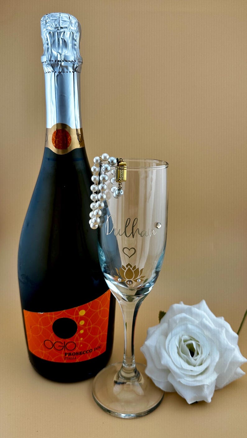 May include: A champagne bottle and a personalized champagne flute with the word "Dulhan" and a lotus flower design. A pearl bracelet is draped over the glass, and a white rose is in the background.