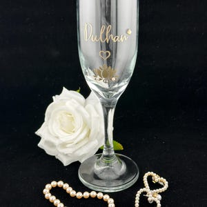 May include: A clear champagne flute with gold lettering that reads "Dulhan", a heart, and a lotus flower design. A white rose, a pearl bracelet, and a pearl heart-shaped brooch are also present.