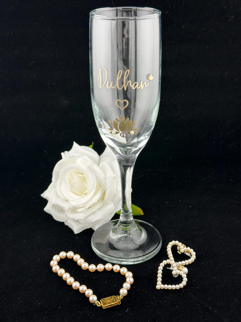 May include: A clear champagne flute with gold lettering that reads "Dulhan", a heart, and a lotus flower design. A white rose, a pearl bracelet, and a pearl heart-shaped brooch are also present.