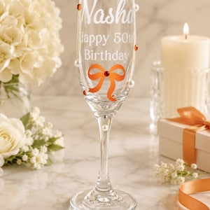 May include: A clear glass champagne flute featuring the white etched text Nasha Happy 50th Birthday. The glass is decorated with an orange bow graphic, small white pearls, and orange and silver rhinestones applied to the exterior surface.